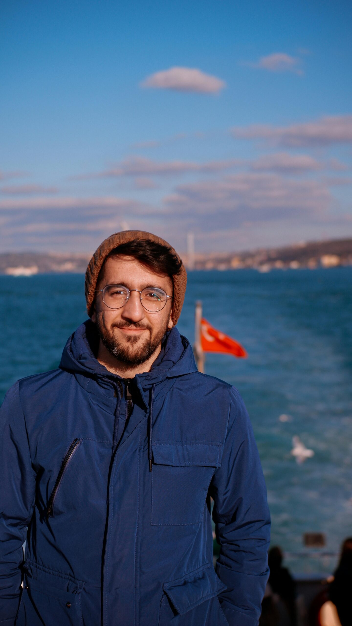 A man in a blue jacket stands by the Bosporus in Istanbul with a view of the sea and Turkish flag.